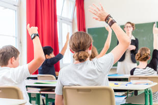 Bild vergrößern: School class teacher giving lesson in front of a blackboard or board teaching students or pupils, they are raising their hands as they know all the answers